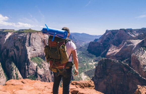 A person with a backpack and a blue rolled-up sleeping mat standing on the edge of a canyon, overlooking a river and tall cliffs under a partly cloudy sky.