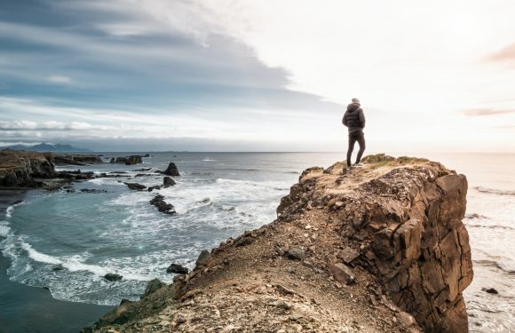 A person in a dark jacket and pants stands on a rocky cliff overlooking the ocean at sunset, with clouds and waves crashing below.