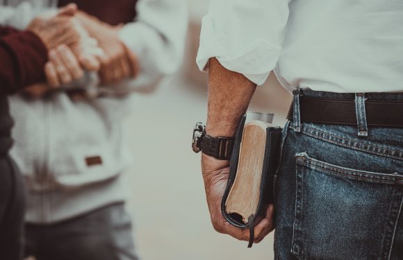 A person in a white shirt holding a Bible, standing next to another person in a gray hoodie and black pants, holding a mug.