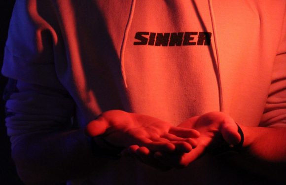 A person wearing a hoodie with the word "SINNER" in bold black letters on the chest, illuminated by red and blue lighting in a dark environment.