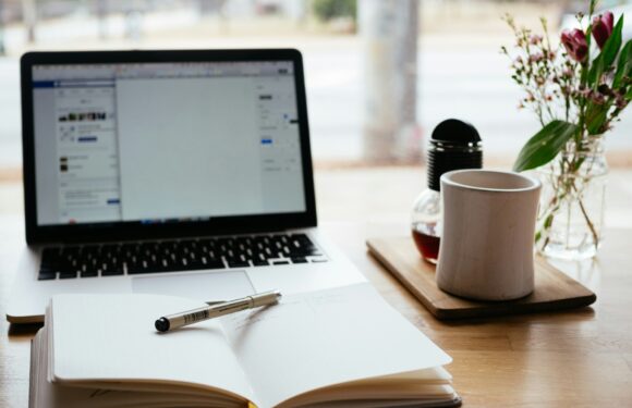 A workspace with an open notebook and pen on a wooden desk, a laptop displaying a social media page, a flowered vase, a coffee mug, and a jar with tea.