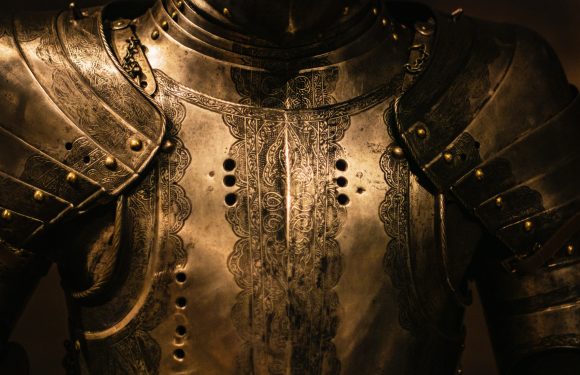 A close-up of an ornate, engraved suit of armor covering the shoulders and chest, with detailed floral and scroll patterns etched into the metal.