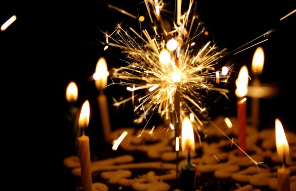 A lit sparkler creates bright sparks and a glowing light, with candles surrounding it on a dark background.