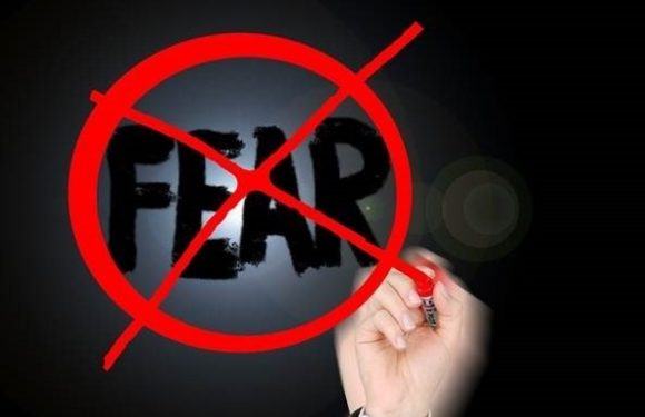 A hand holding a red marker drawing a red circle with a diagonal line over the word "FEAR" on a black background, symbolizing opposition to fear.