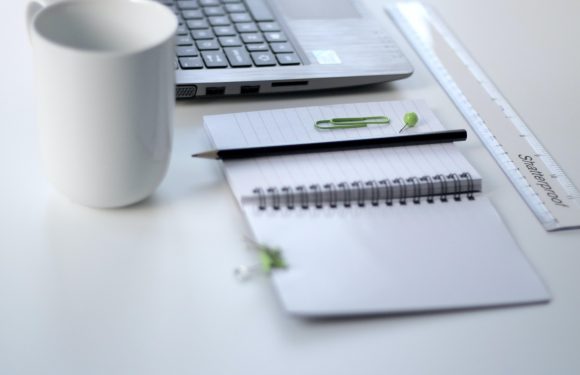 A white desk with a notepad, black pen, green paperclip, small green push pin, laptop, ruler, and a white mug, all arranged neatly with a clean, minimal aesthetic.