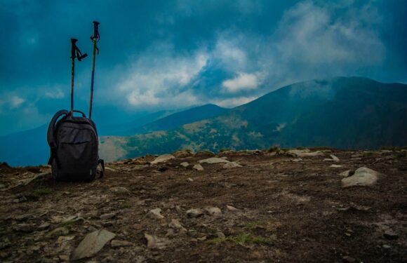 A black backpack with two trekking poles stuck into it, set on rocky ground with mountains and cloudy sky in the background.