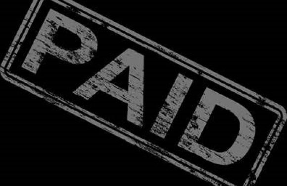 A person in a uniform and beard stands outside next to a lawn mower, holding a snow shovel, with a fence and trees in the background.