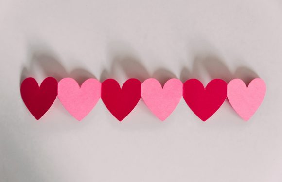 Six paper hearts, alternating red and pink, aligned in a row on a white background with their shadows cast behind them.
