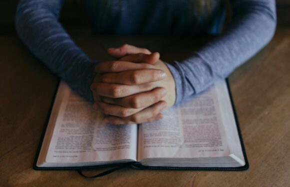 A person with clasped hands resting on an open Bible on a wooden table. The individual is wearing a long-sleeved, blue-gray shirt.