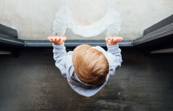A child with blonde hair, wearing a light gray hoodie, looks out a glass door, with their hands pressed against the glass. Their reflection is visible on the door.