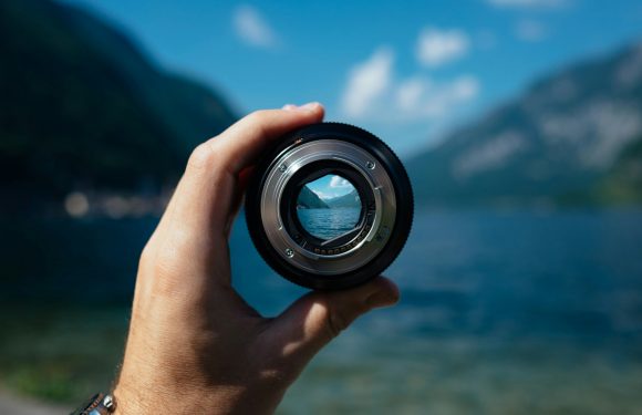A hand holds a camera lens, capturing a scenic view of a lake with mountains and clouds in the background, seen through the lens.