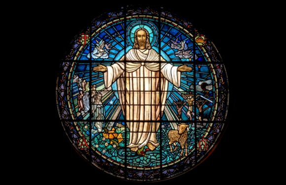 A stained glass window depicting Jesus Christ with open arms, surrounded by angels, a lamb, and various religious symbols, set against a dark background.