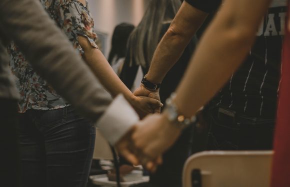 People holding hands in a line, standing close together indoors, some wearing patterned or striped clothing, in a gesture of unity or support.