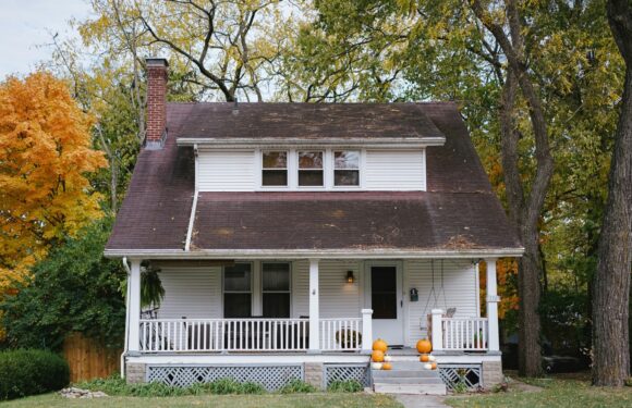 A white two-story house with a front porch decorated with pumpkins, surrounded by trees with autumn-colored leaves, and a chimney on the left side.