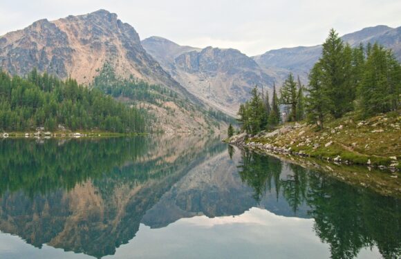 A tranquil mountain lake reflecting rugged peaks, dense green pine trees, and a cloudy sky in a scenic wilderness area.