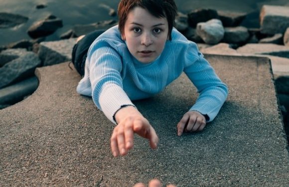 A young person with short dark hair, wearing a blue and white sweater, is reaching out to an outstretched hand from the edge of a body of water, with rocks nearby.