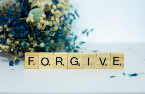 Scrabble tiles spelling "FORGIVE" placed on a white surface with dried flowers and greenery in the background.