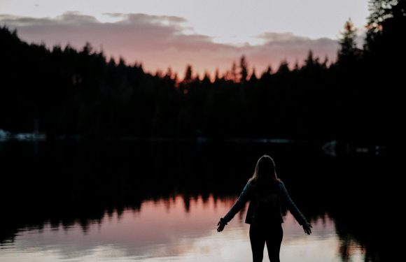 A person with long hair stands near a calm body of water during sunset, arms outstretched, with a silhouette of trees and a colorful sky in the background.