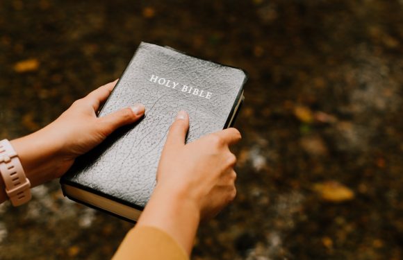 Hands holding a black leather Holy Bible against a blurred outdoor background with scattered leaves. One hand has a light pink watch, and the other wears a yellow sleeve.