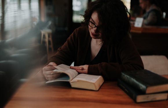 A woman with curly dark hair and glasses sits at a wooden table, reading a book in a cozy, dimly-lit cafe with a blurred background.
