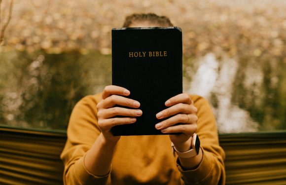 Person holding a Holy Bible in front of their face, wearing a mustard-colored long-sleeve shirt and smartwatch, with a blurred outdoor background.