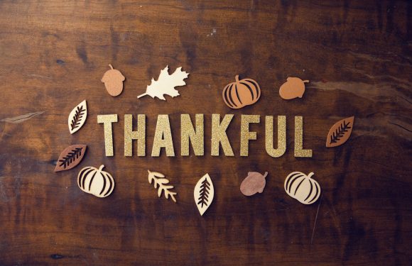 "Thankful" spelled out in glittery gold letters on a wooden surface, surrounded by paper cutouts of pumpkins, acorns, and leaves in warm fall tones.