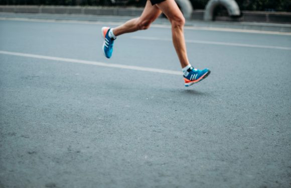 Runner wearing blue shoes and shorts, sprinting on an outdoor paved track, with a row of tires visible in the background.