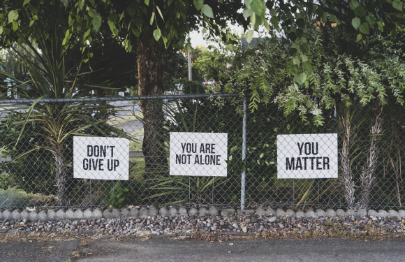 Three signs attached to a chain-link fence with greenery and trees in the background. The signs say: "DON'T GIVE UP", "YOU ARE NOT ALONE", and "YOU MATTER".