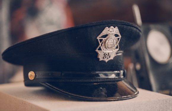 A black police hat with a silver badge featuring wings and the number 86, resting on a beige surface. A piece of equipment is blurred in the background.