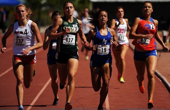 Six female runners participating in a race on a track, wearing athletic uniforms with race numbers, with a sunny outdoor setting.