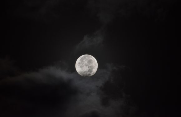 A bright full moon illuminated the dark night sky, partially obscured by clouds. The moon's surface details are clearly visible.