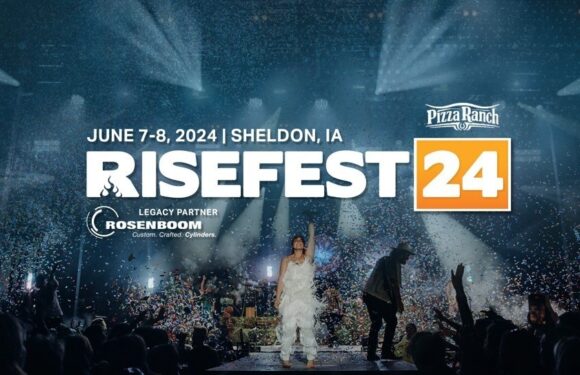 A woman in a white dress stands on a stage with her arm raised amid colorful confetti, with several people nearby, under bright stage lights.