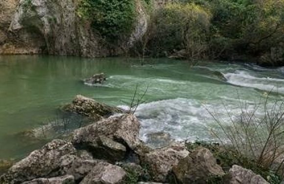 A river with turquoise water flows over rocks, surrounded by lush green trees and rocks along the riverbank.