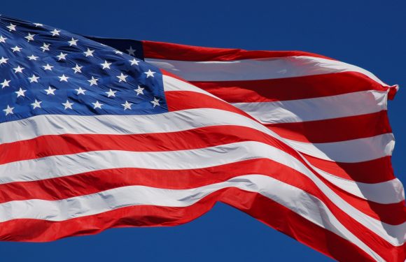A waving American flag with red and white stripes and white stars on a blue field, set against a clear blue sky.