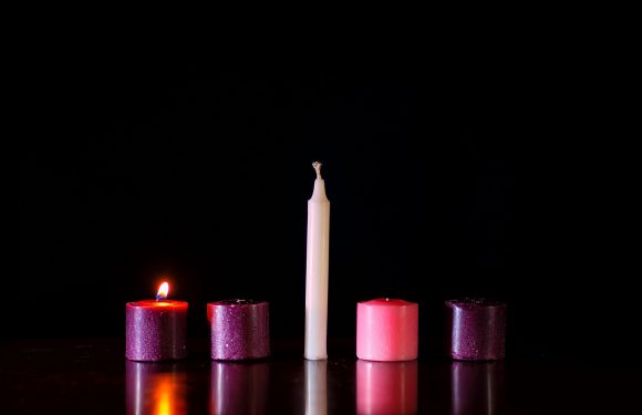 Four candles, with the second one from the left lit, are arranged on a reflective surface against a black background. The candles are pink, purple, and white.