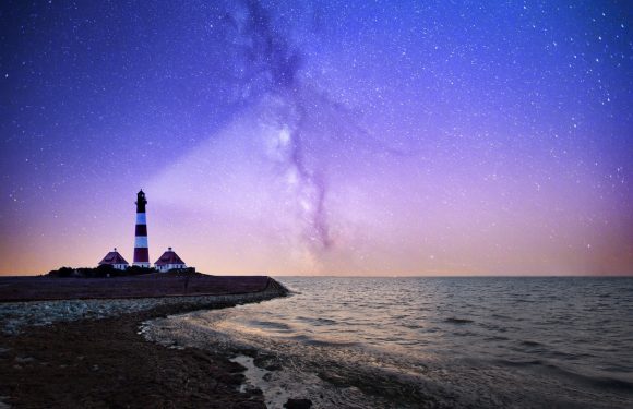 A lighthouse on a small landmass by the water under a vivid, star-filled night sky with the Milky Way galaxy visible.