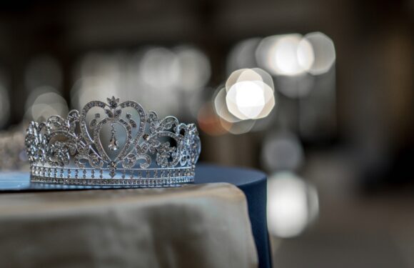 A sparkling silver tiara with intricate designs and gemstones rests on a surface with blurry lights in the background.
