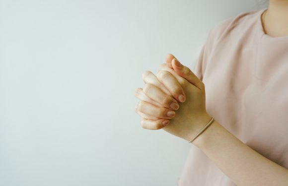 A person with folded hands wearing a light pink shirt and a neutral-colored bracelet against a plain light background.