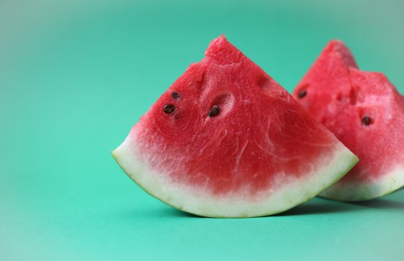 Two slices of watermelon with a red interior and black seeds sit on a teal background. The slices are fresh and juicy, with a green rind at the base.