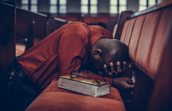 A man in an orange uniform with black hair, leaning forward with his head on a pew, appears exhausted or overwhelmed, with a large book and reading glasses placed nearby.