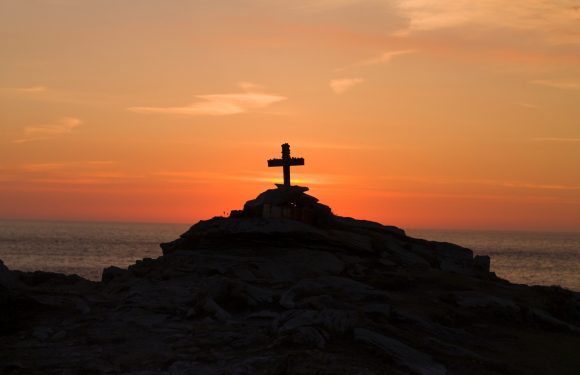 A silhouette of a cross on a rocky hilltop at sunset over the ocean, with the sky painted in orange and pink hues.