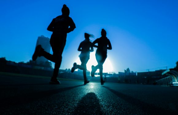 Silhouettes of three runners running on a track during sunset, with a bright sun behind them and a building in the distance.