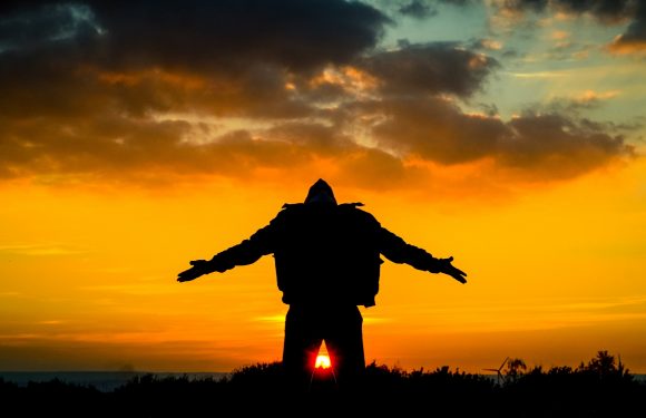 A person standing outdoors with arms outstretched at sunset, wearing a jacket and a backpack, with a vibrant sky and clouds overhead.