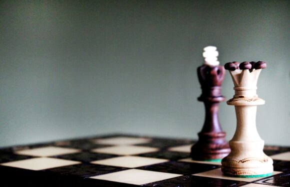A white chess king and a darker brown chess king stand on a black-and-white checkered chessboard with a plain, muted green background.