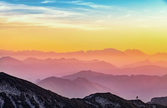 A lone hiker stands on a rocky mountain ridge at sunset, overlooking multiple mountain ranges with colorful, layered sky in shades of blue, pink, orange, and yellow.