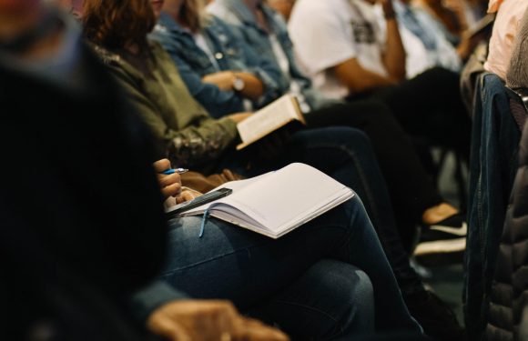 People seated in rows, some with notebooks and pens, attending a presentation or lecture in a dimly lit room.