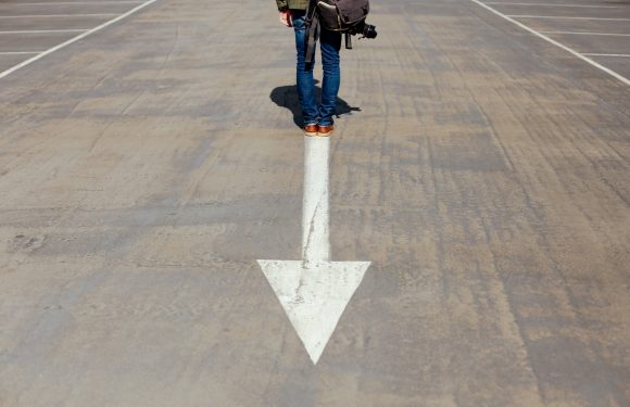 A person standing on a parking lot with a white downward arrow painted on the ground, holding a camera and wearing a green jacket, blue jeans, and brown shoes.