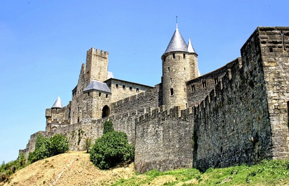 A historic stone castle with turrets and towers is situated on a grassy hill under a clear blue sky. The castle features high stone walls and narrow windows.