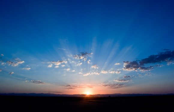 A person in a Pizza Ranch uniform holding a tray of food in front of a sunset sky with rays of sunlight and scattered clouds.