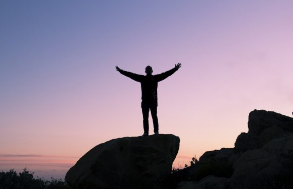 A person stands with arms outstretched on a large rock formation during sunset, silhouetted against a pastel pink and purple sky.